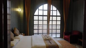 A hotel room with a large arched window, a bed with white linens, a red armchair, and a small table with a lamp.