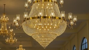 A grand chandelier with multiple tiers and candle-like lights, hanging in a spacious room with high ceilings and large windows. - Qila Hotel (Moradabad)