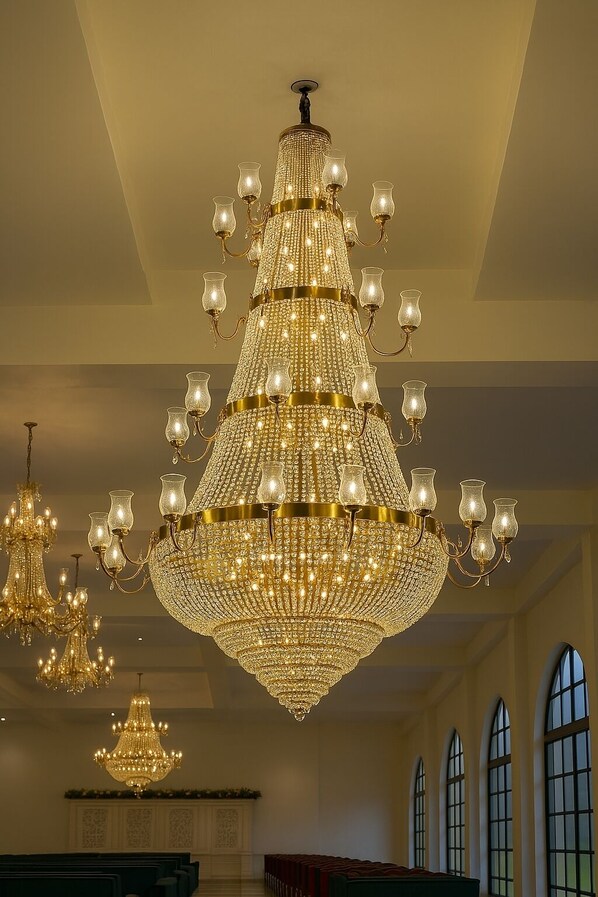 A grand chandelier with multiple tiers and candle-like lights, hanging in a spacious room with high ceilings and large windows. - Qila Hotel (Moradabad)