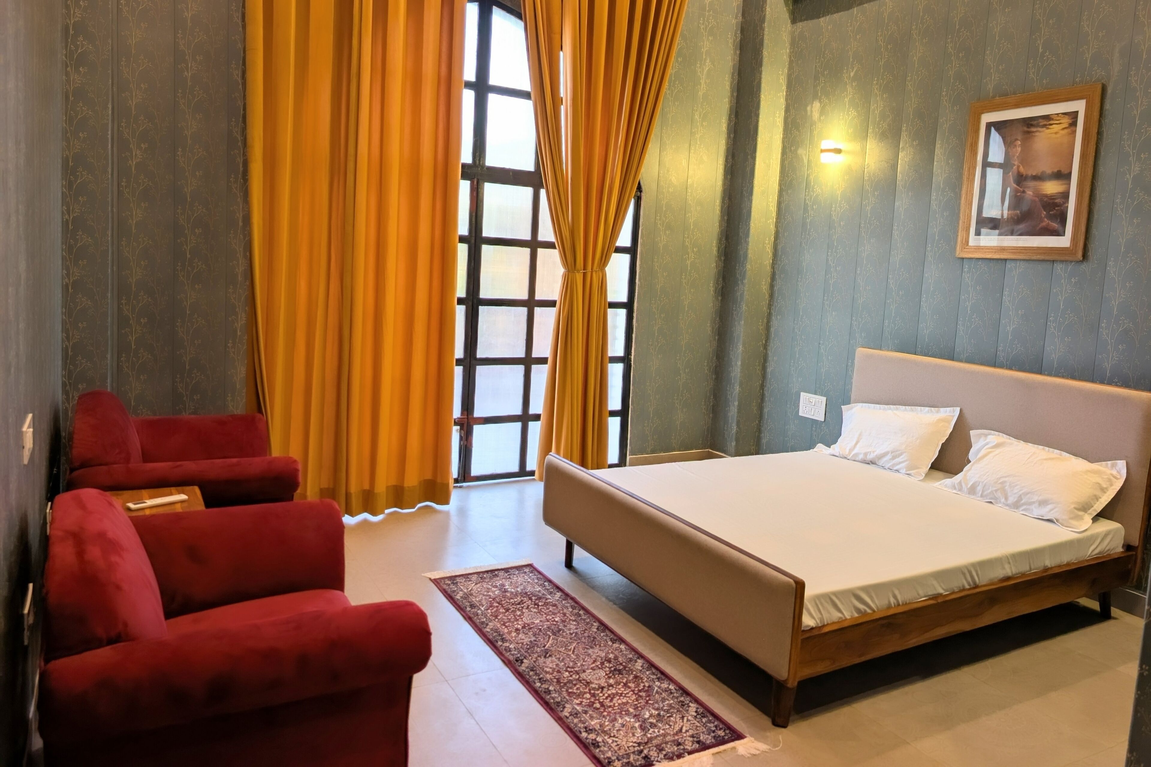 A bedroom with a bed, red chairs, a window with curtains, and a wall-mounted picture.