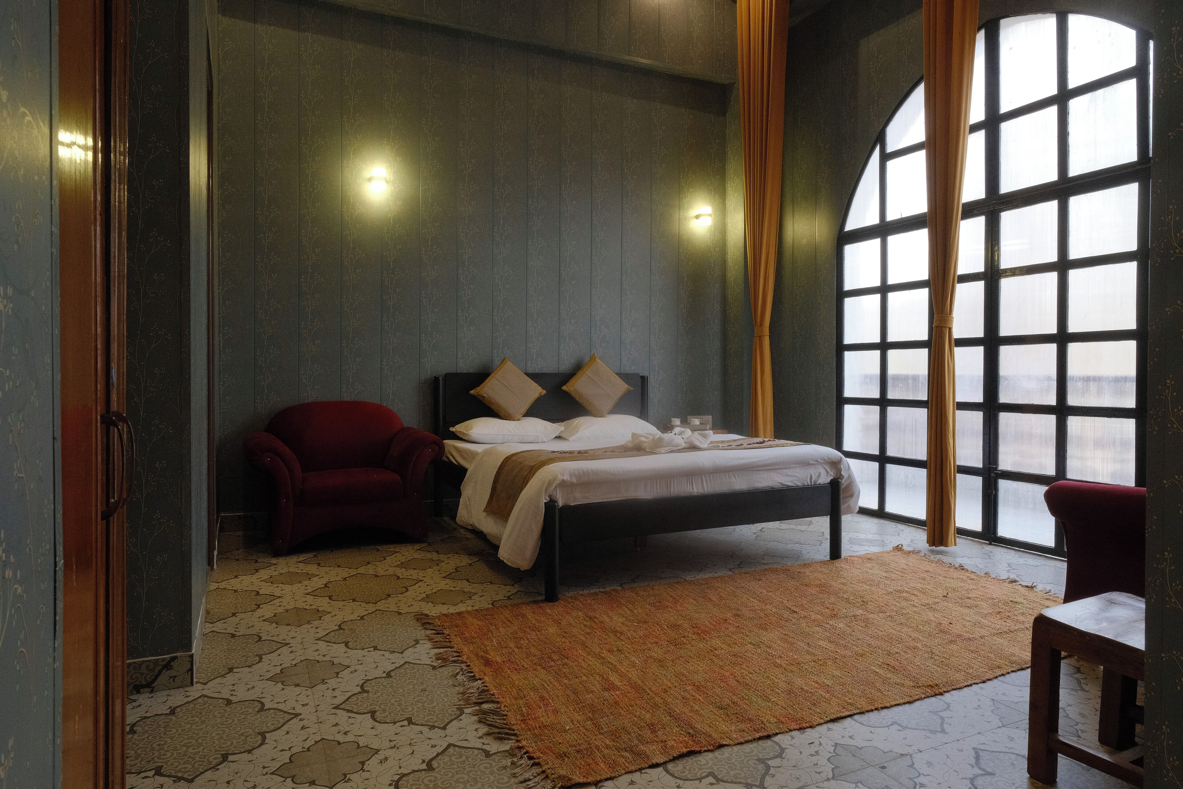 Une chambre à coucher avec un lit, un fauteuil rouge, une grande fenêtre et un tapis à motifs.