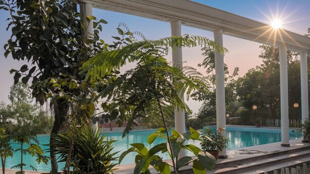 A pool area with a clear view of the pool, surrounded by lush greenery and white columns. - Qila Hotel (Moradabad)