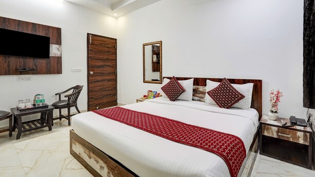 A hotel room with a bed, a TV mounted on the wall, a wooden wardrobe, a small table with a chair, and a nightstand with a phone. - Hotel Amour By RMR Hotels (New Delhi)