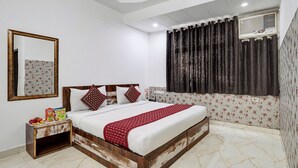 A bedroom with a bed, a nightstand, a mirror, and a window with curtains. - Hotel Amour By RMR Hotels (New Delhi)