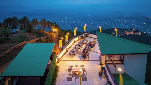 Uma área de refeições ao ar livre com mesas e cadeiras, um braseiro e vista para um horizonte urbano ao entardecer.