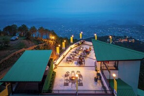 An outdoor dining area with tables and chairs, a fire pit, and a view of a cityscape at dusk. - The Hosteller Dharamshala, Mountain Escape (Dharamshala)