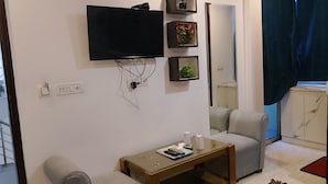 A living room with a flat-screen TV mounted on the wall, a small wooden coffee table, two grey armchairs, and a mirror. - Hotel The Raj Palace (New Delhi)