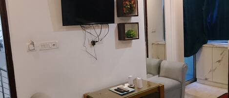 A living room with a flat-screen TV mounted on the wall, a small wooden coffee table, two grey armchairs, and a mirror.