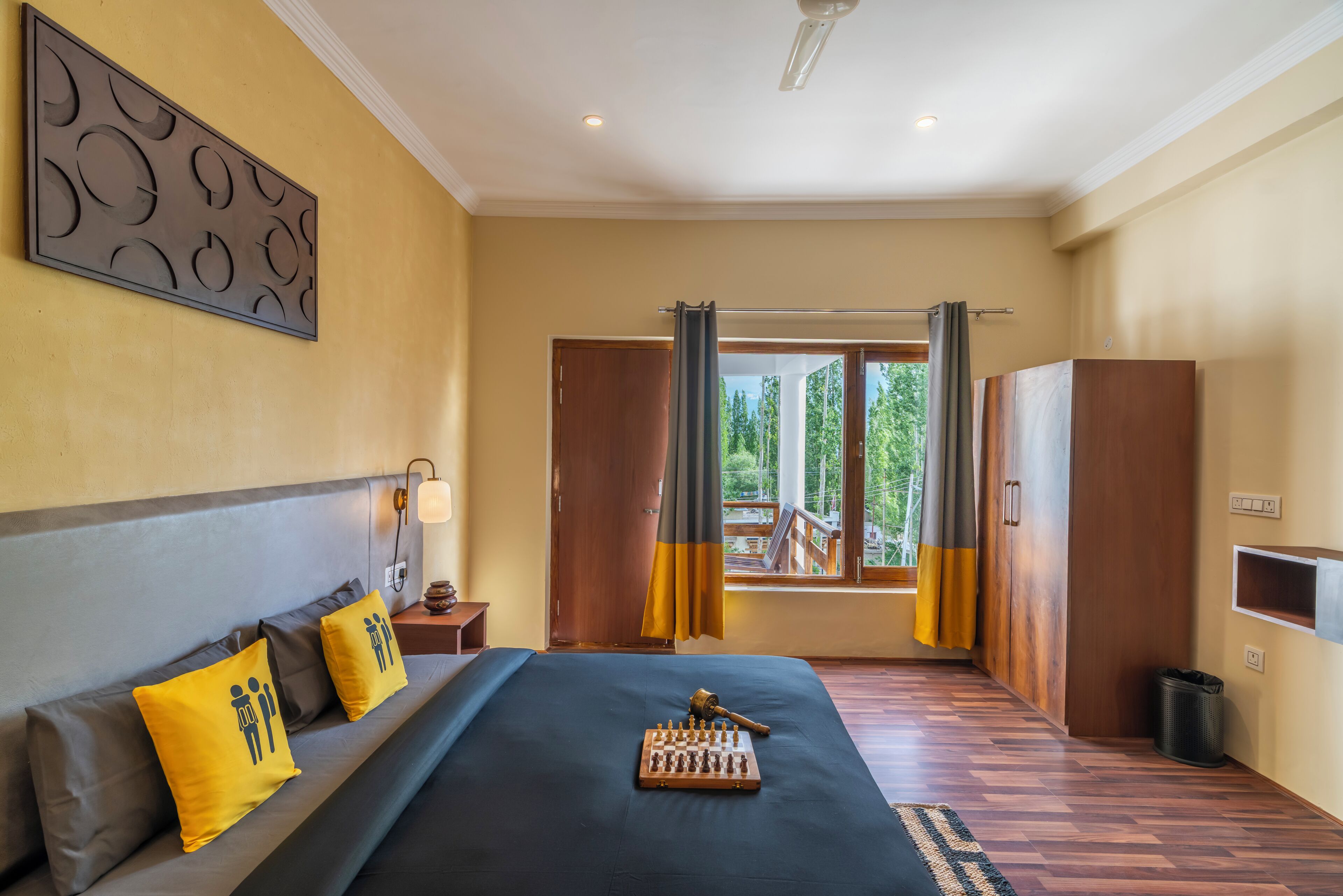 A hotel room with a bed, a chessboard, a nightstand, a lamp, and a view of the outdoors through a window.