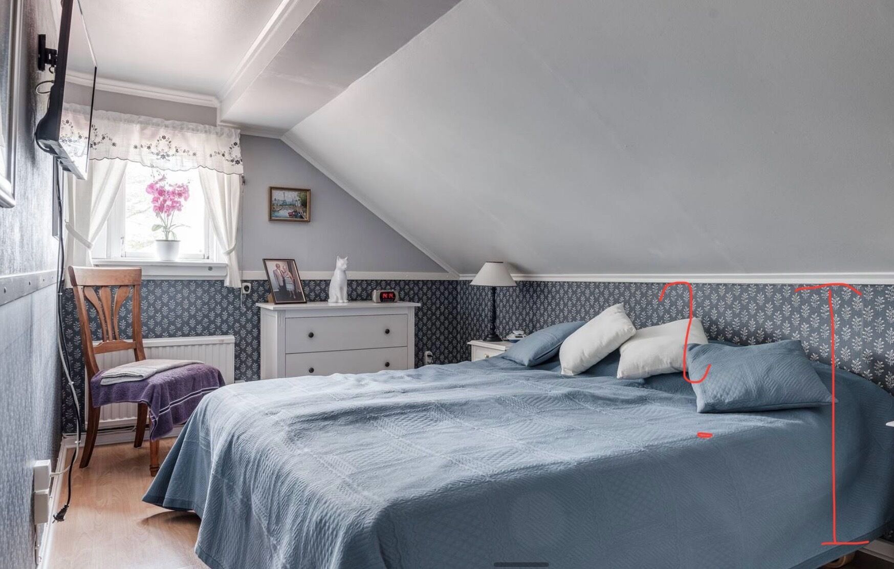 A bedroom with a sloped ceiling, a bed with blue bedding, a nightstand, a chair, a dresser, a window with curtains, and a wall with a patterned wallpaper.
