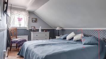 A bedroom with a sloped ceiling, a bed with blue bedding, a nightstand, a chair, a dresser, a window with curtains, and a wall with a patterned wallpaper.