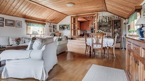 A spacious living area with a high wooden ceiling, a dining table with chairs, and a kitchen area visible through an open doorway. - A house by the forest with a stream and plenty of peace and quiet (Stenis)