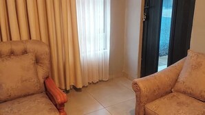 Ein Zimmer mit zwei beigen Sesseln, einem Holzstuhl, einem Fenster mit transparenten Vorhängen und einer Tür, die nach draußen führt.