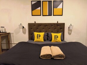 Une chambre d’hôtel avec une tête de lit capitonnée, deux lampes de chevet, un lit recouvert d’une couvre-lit sombre, deux oreillers ornés d’un motif jaune et trois œuvres d’art encadrées accrochées au mur.