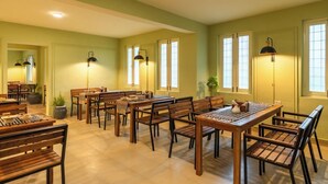 Zona pranzo con diversi tavoli e sedie in legno, illuminazione a incasso nel soffitto e ampie finestre con infissi bianchi.
