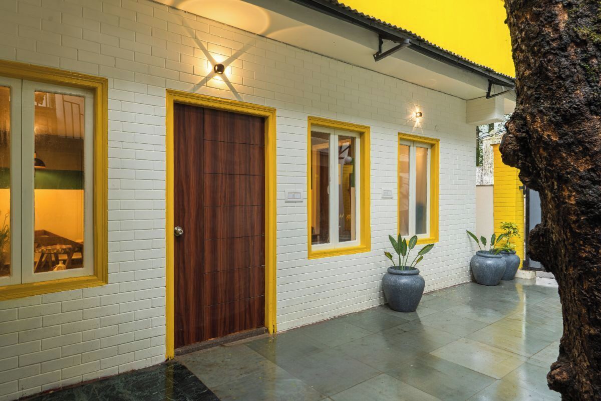 A building with white brick walls and yellow trim around windows and doors. There are potted plants on the ground.