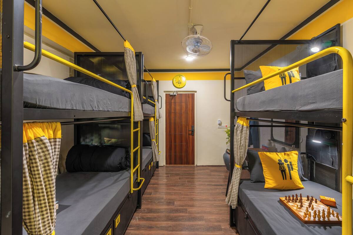 A dormitory room with bunk beds, a fan, a clock, and a chessboard.