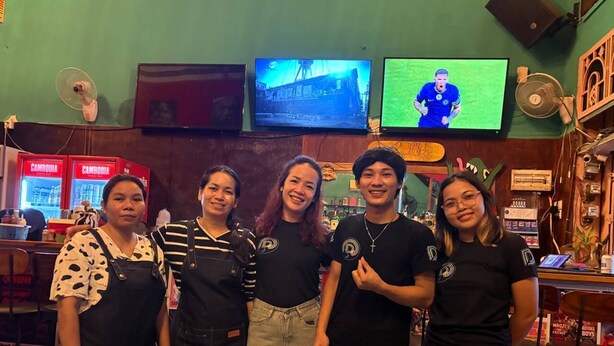 A group of people standing in a bar area with televisions displaying sports events. - The Deck Toul Tompoung (Phnom Penh)