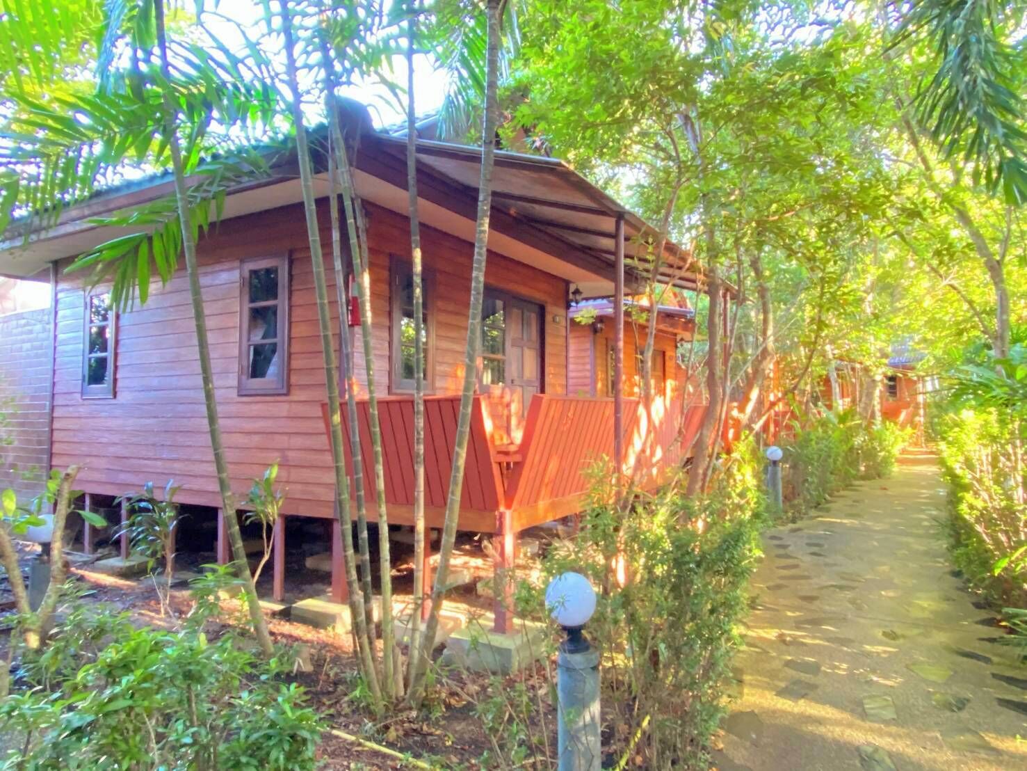 A rustic wooden house surrounded by dense greenery, with a clear path leading to a small structure in the distance.