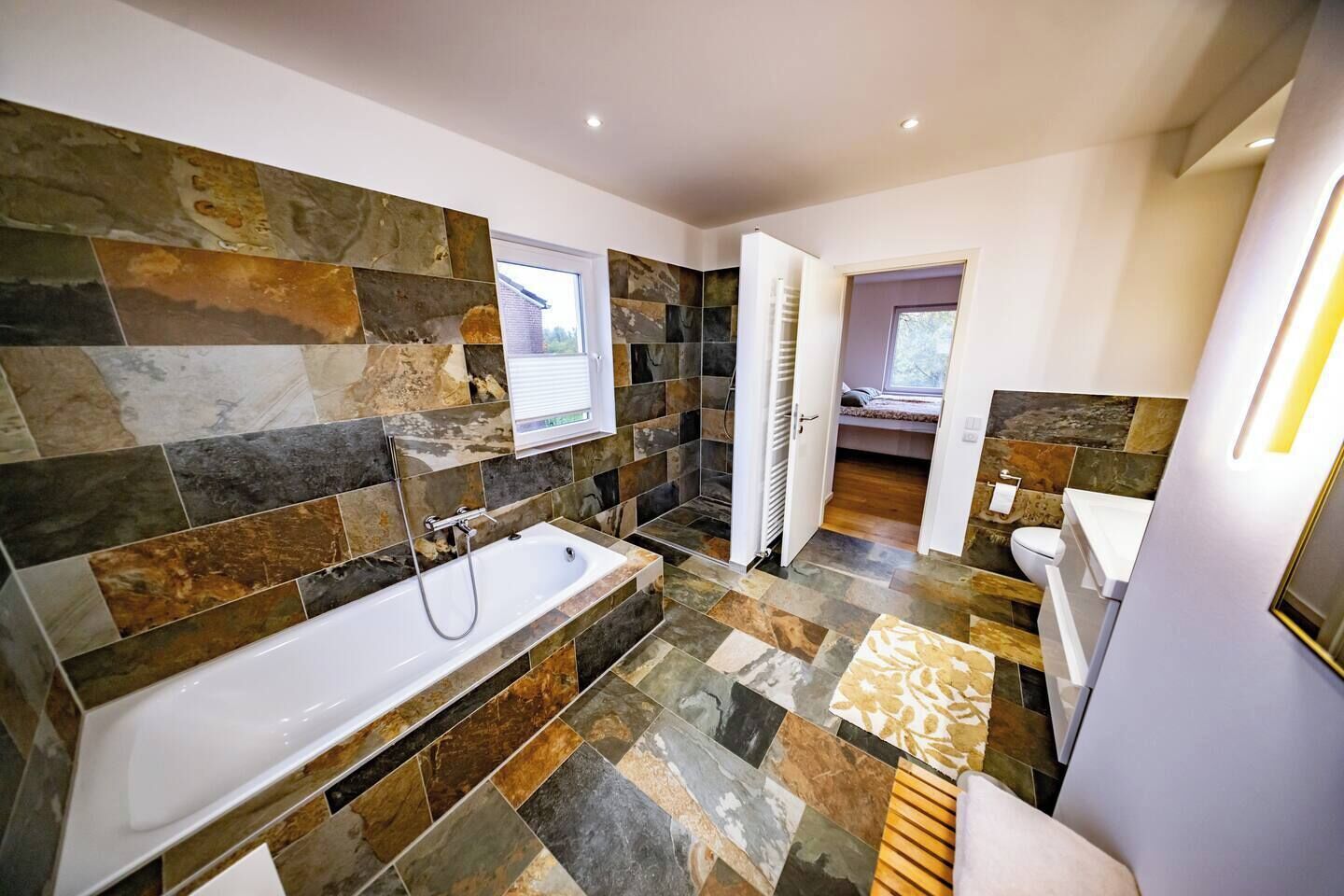 A modern bathroom with a large bathtub, a shower, and a toilet. The walls are tiled with various stone patterns.