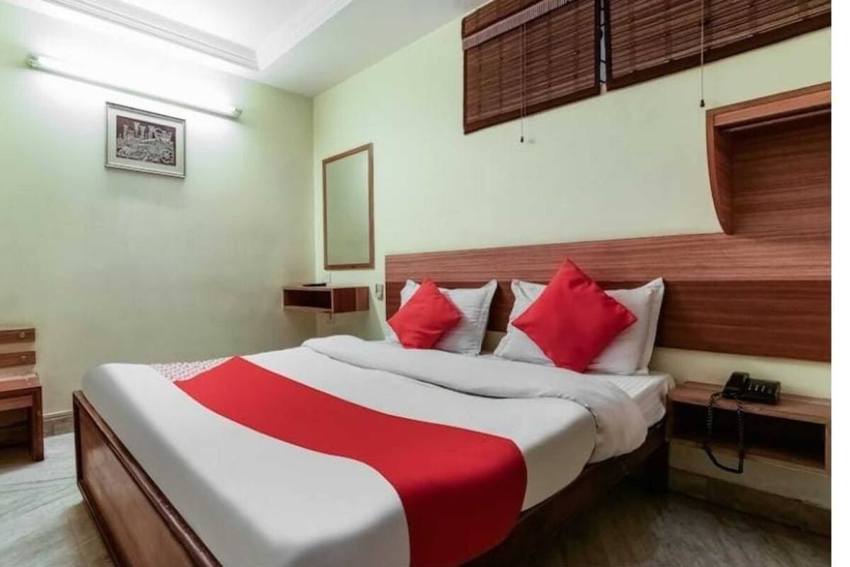A hotel room with a bed, a bedside table, a nightstand, a mirror, and a window with blinds.