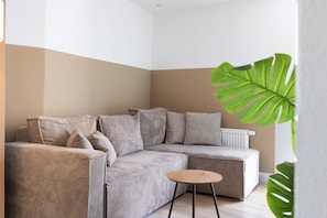 A corner living room with a beige sofa, a small round table, and a large green plant.