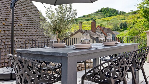 Un espacio de comedor al aire libre con una mesa, sillas, una valla metálica y vistas a una colina.