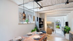 Un comedor con una mesa de madera dispuesta para cuatro, una lámpara colgante y vistas a una sala de estar con un sofá y plantas.