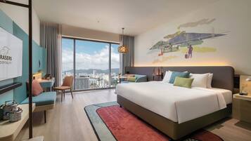 A modern hotel room with a large bed, a desk, a chair, and a view of a cityscape through large windows.