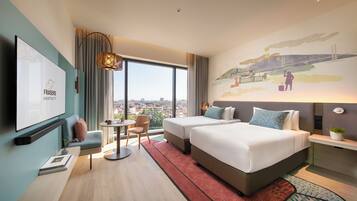 A hotel room with two beds, a TV, a desk, a chair, and a large window with a city view.
