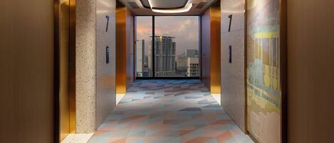 A modern elevator interior with a geometric patterned floor, a cityscape view through the windows, and a ceiling with recessed lighting.
