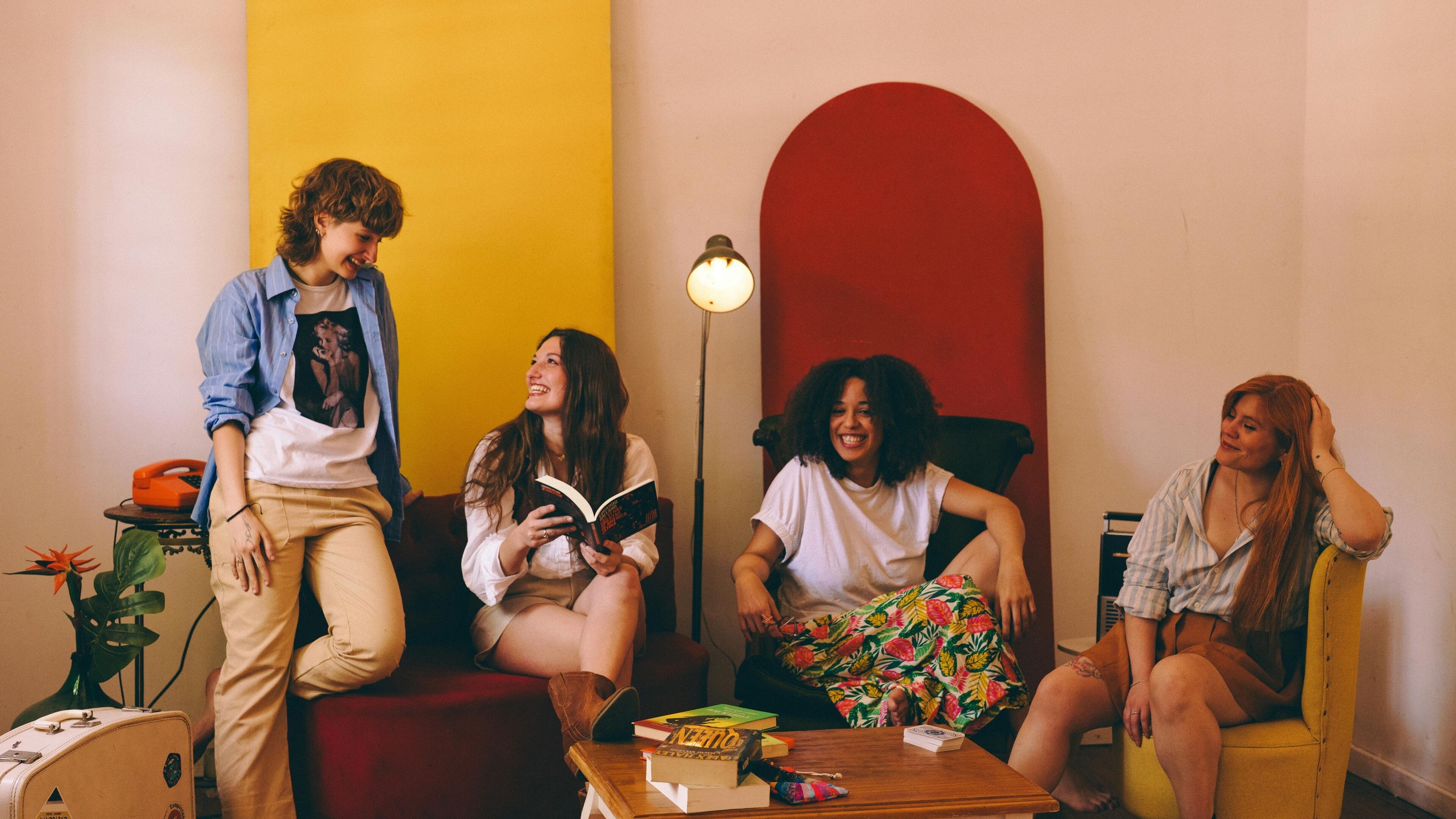 Un grupo de personas sentadas y de pie en una habitación con un arco rojo, una lámpara y una mesa con libros y una maleta.