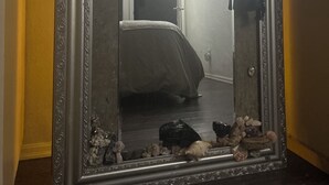 A mirror with an ornate frame reflecting a room with a bed, a door, and a wall-mounted light fixture.