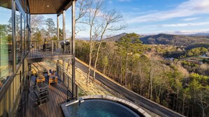 Ein modernes Gebäude mit Glasfassade, einer Holzterrasse und Sitzgelegenheiten im Freien sowie Blick auf ein bewaldetes Tal.
