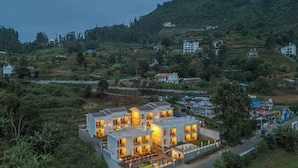 Un immeuble résidentiel moderne à plusieurs étages, doté de grandes fenêtres et de balcons, entouré d’une végétation luxuriante et de collines.