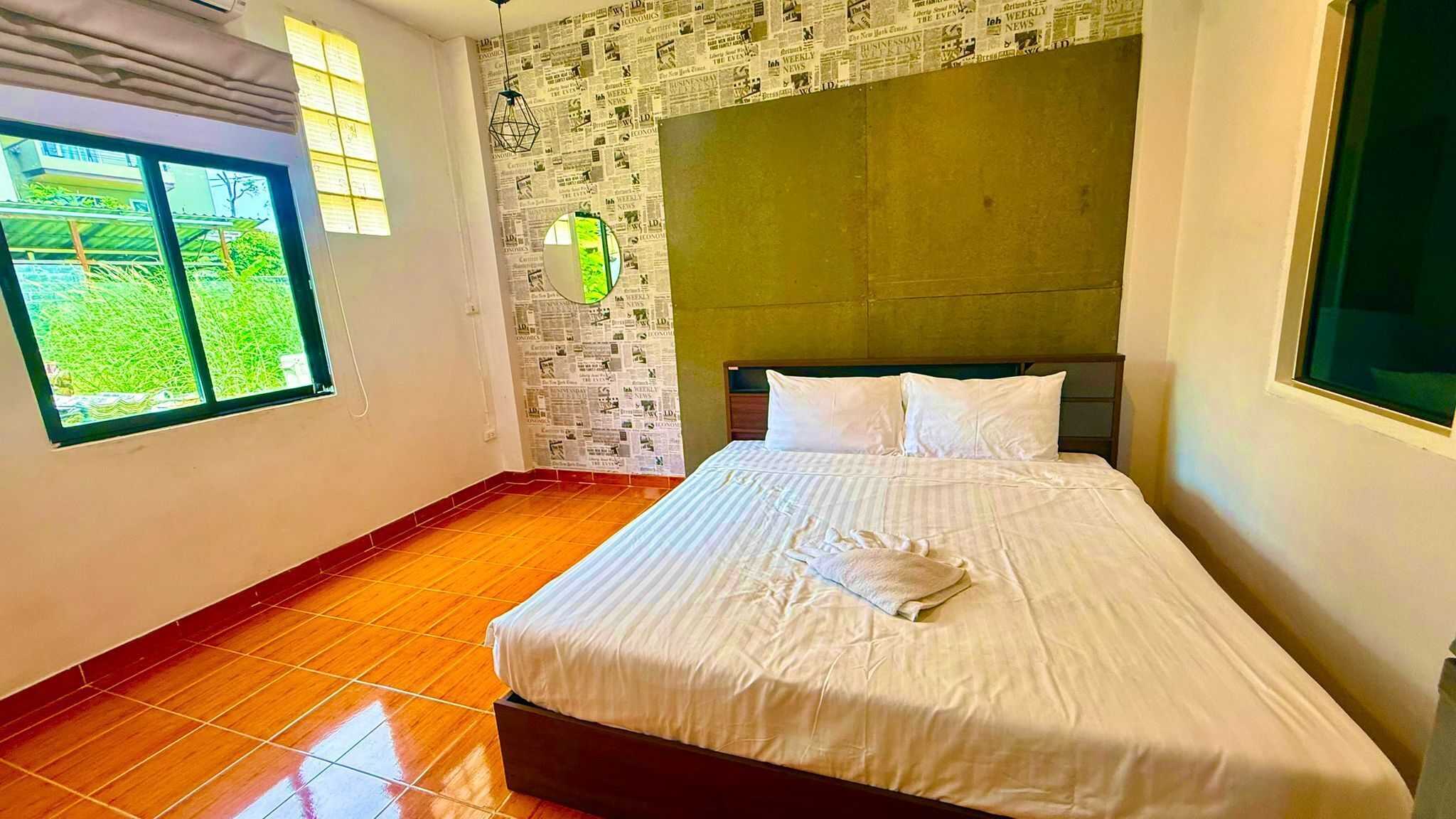 A hotel room with a bed, a window with a view of greenery, and a wall covered with newspaper clippings.