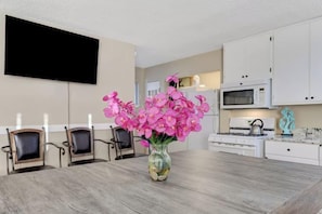A dining area with a wooden table, chairs, a vase of flowers, a flat-screen TV, and white cabinets. - Strip-Side Villa, 6 Beds, Pool, Mini Golf, Karaoke (Las Vegas)