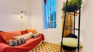 Una stanza piccola e accogliente con pavimento a scacchi, un divano rosso con cuscini decorati, una finestra con vista e una mensola in metallo nero con una pianta.