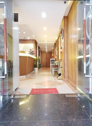 Zona de recepción de hotel con mostrador, felpudo con la palabra "WELCOME" y puerta de cristal de entrada.