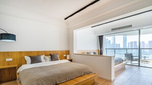 Une chambre moderne avec un grand lit, une tête de lit en bois et une vue sur la ville grâce à de grandes fenêtres.