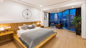 Une chambre moderne avec un grand lit, une tête de lit en bois, des tables de chevet et une vue sur la ville grâce à de grandes fenêtres.