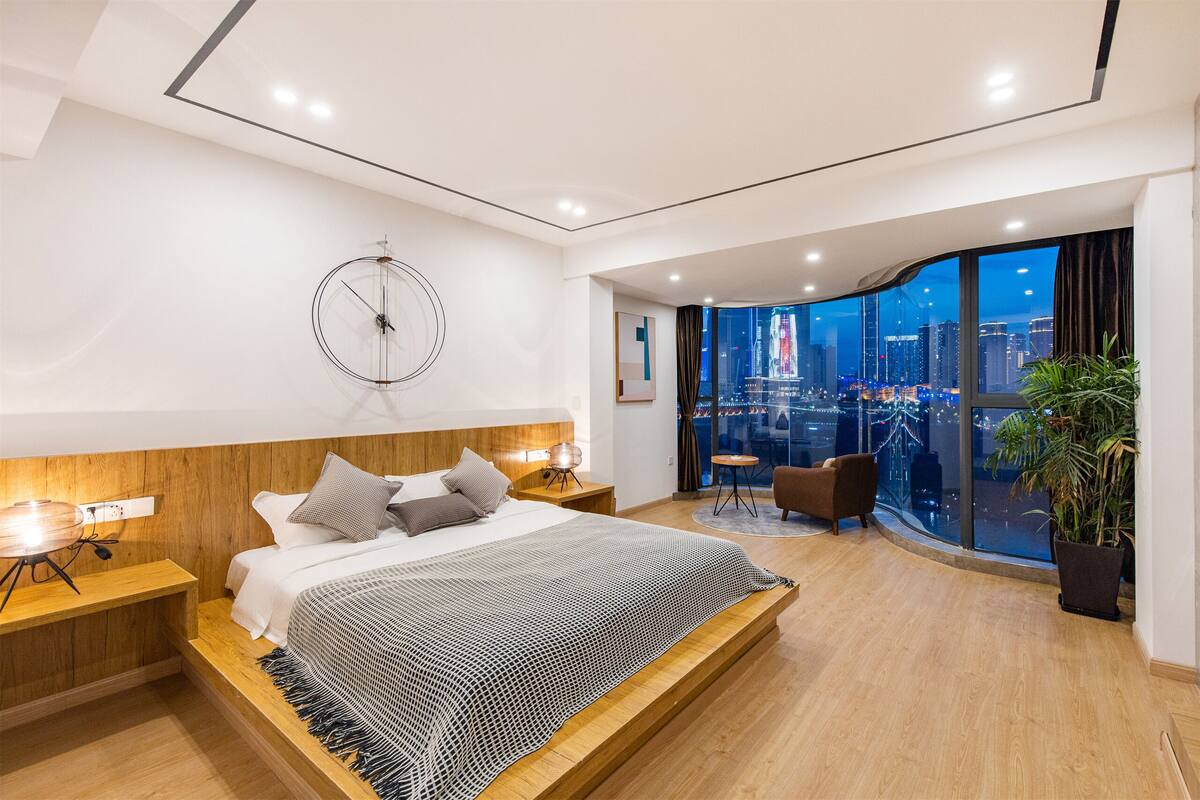 A modern bedroom with a large bed, wooden headboard, bedside tables, and a city view through large windows.