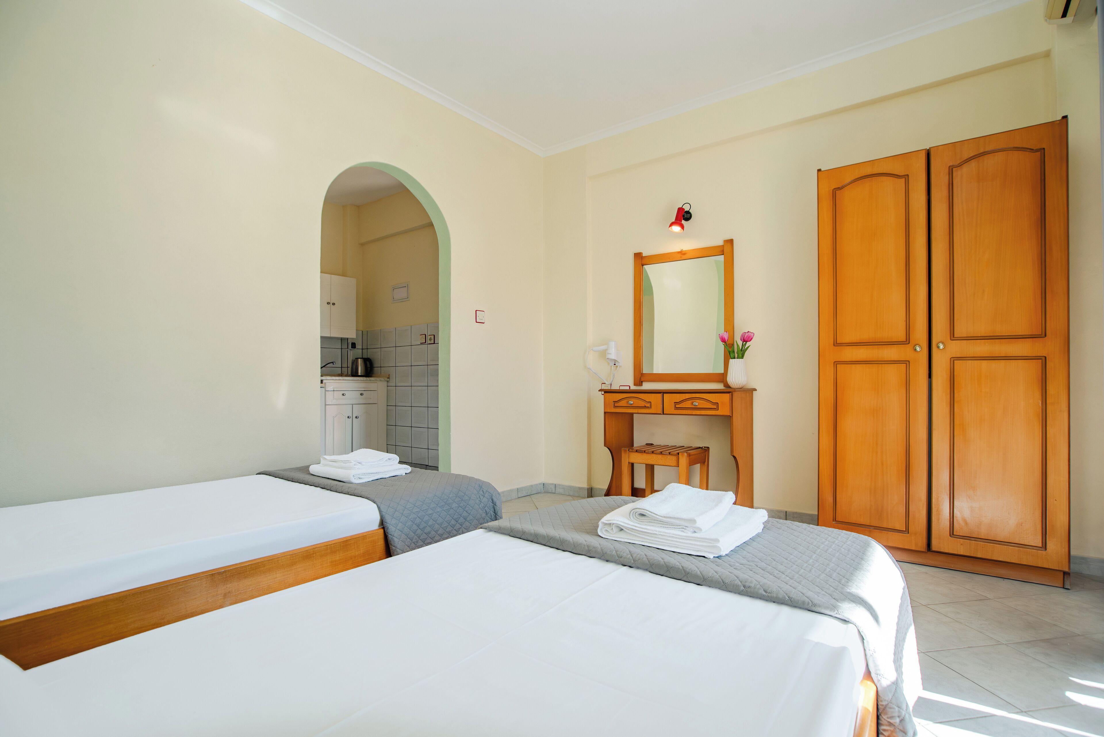 Habitación de hotel con dos camas, un armario de madera, un pequeño escritorio con espejo y una kitchenette al fondo.