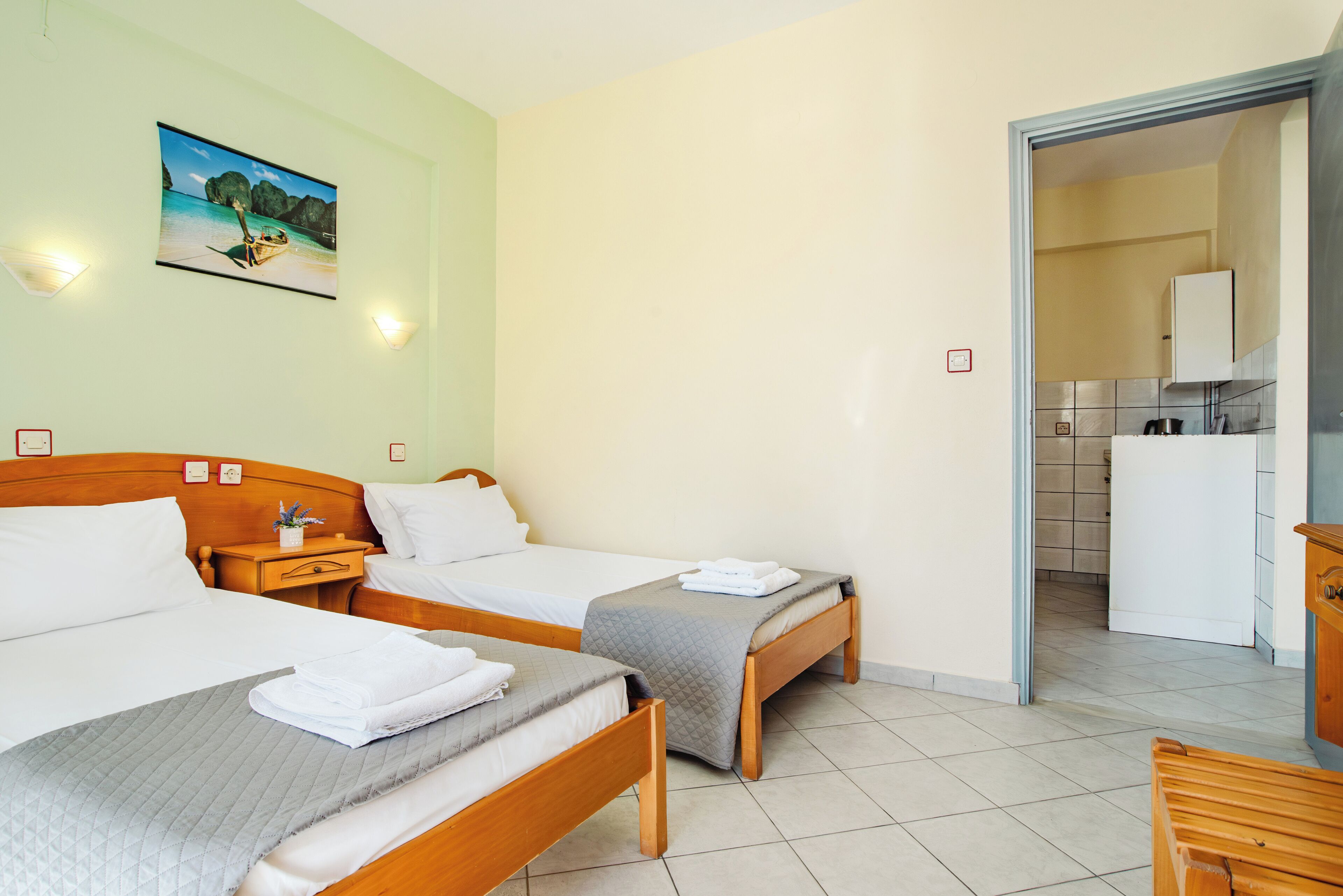 A hotel room with two beds, a painting of a beach scene, a kitchenette, and a bathroom visible through an open door.