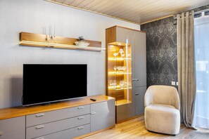 Un soggiorno moderno con TV a schermo piatto, un mobiletto in legno con vetrate, una poltrona beige e carta da parati decorata.