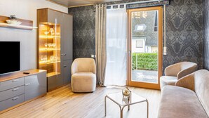 A modern living room with a flat-screen TV, a glass cabinet with wine glasses, a beige armchair, a sofa, and a glass coffee table. - Apartment 'Marisa's Sauerland Dream' with Shared Terrace, Garden & Wi-Fi (Brilon)