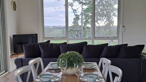 A dining area with a wooden table set for four, surrounded by white chairs, with a dark sofa and a television in the background. - Grasmere Lodge with Garden (Gruyere)