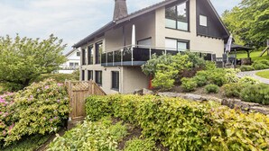 Ein zweistöckiges Haus mit Balkon, umgeben von einem Garten mit verschiedenen Pflanzen und einem Holzzaun.