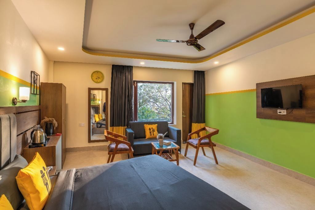 Quarto de hotel com cama, duas poltronas, um sofá, televisão na parede, ventilador de teto e janela com cortinas.