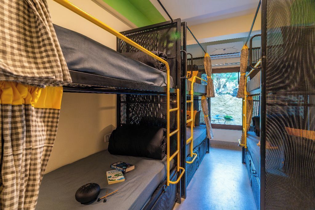 A bunk bed room with a checkered curtain, a small table with a book and a mouse, and a window with a view of trees.